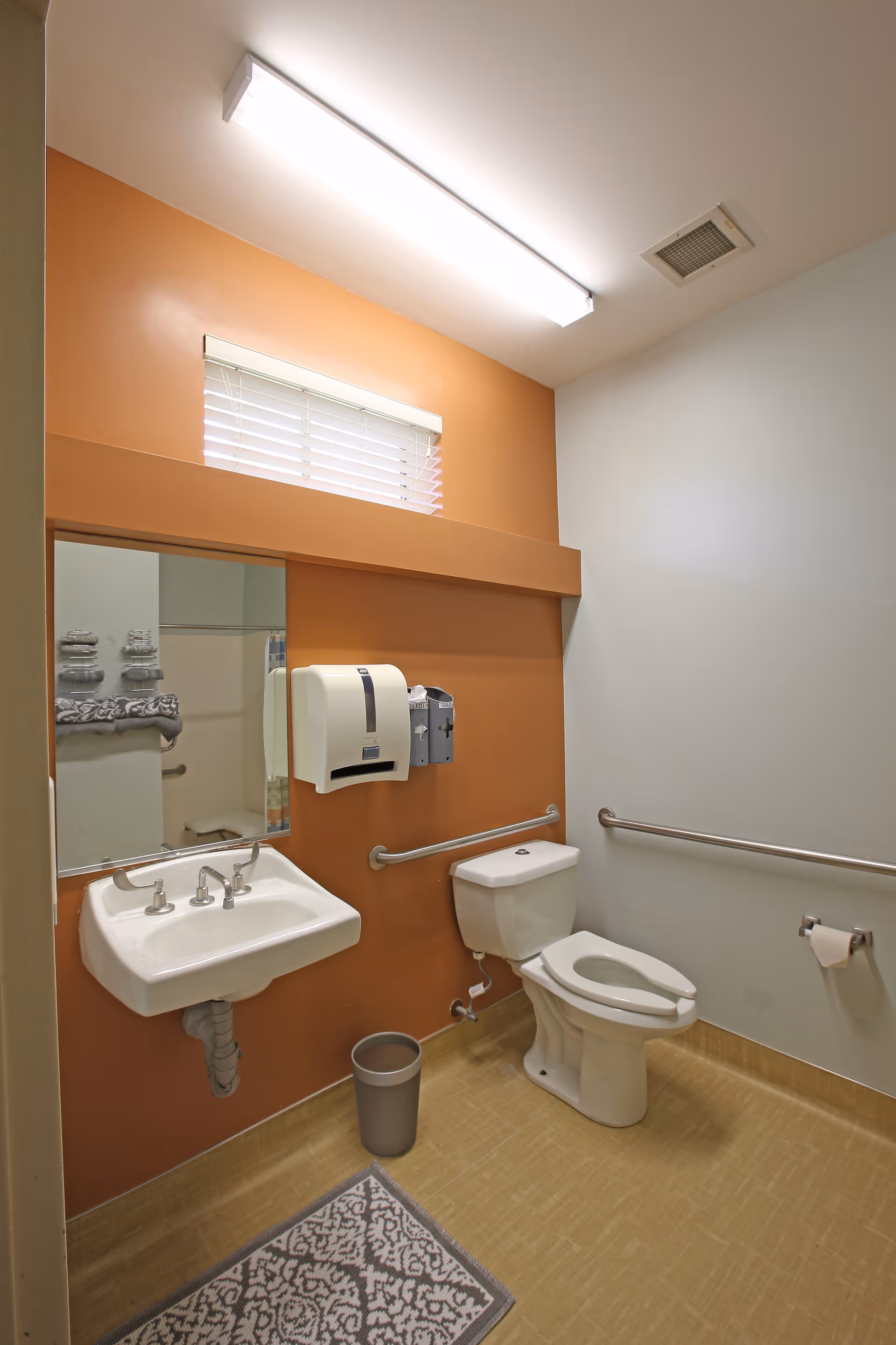 A clean and accessible bathroom with a white toilet, a wall-mounted sink, a large mirror, and grab bars on the walls. The walls are painted orange and white, with a small window above the mirror. There is a paper towel dispenser and a soap dispenser mounted on the wall, a small trash can on the floor, and a patterned rug in front of the sink.