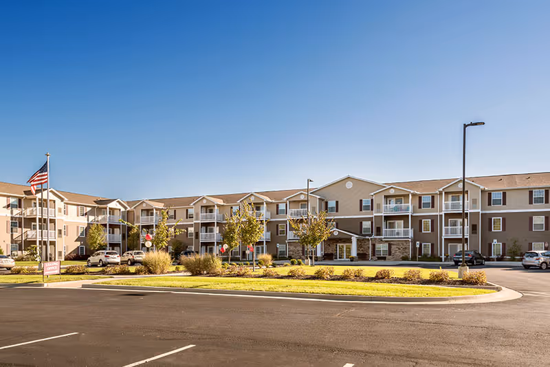 Exterior view of a three-story senior living facility building with balconies, surrounded by a parking lot and landscaped greenery under a clear blue sky.