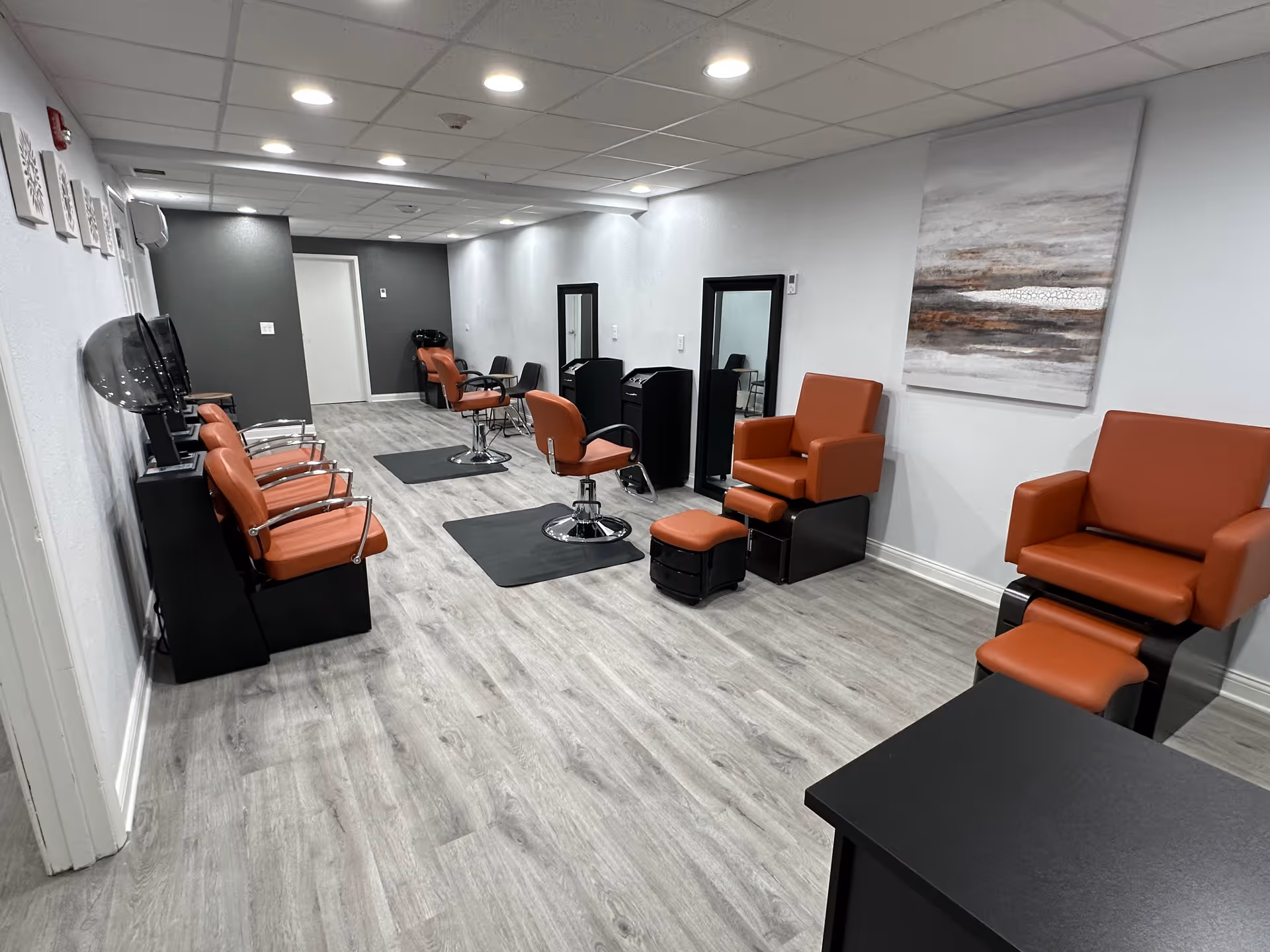 Modern salon-style interior with orange salon chairs, hair stations, and footrests arranged along a light wood floor.