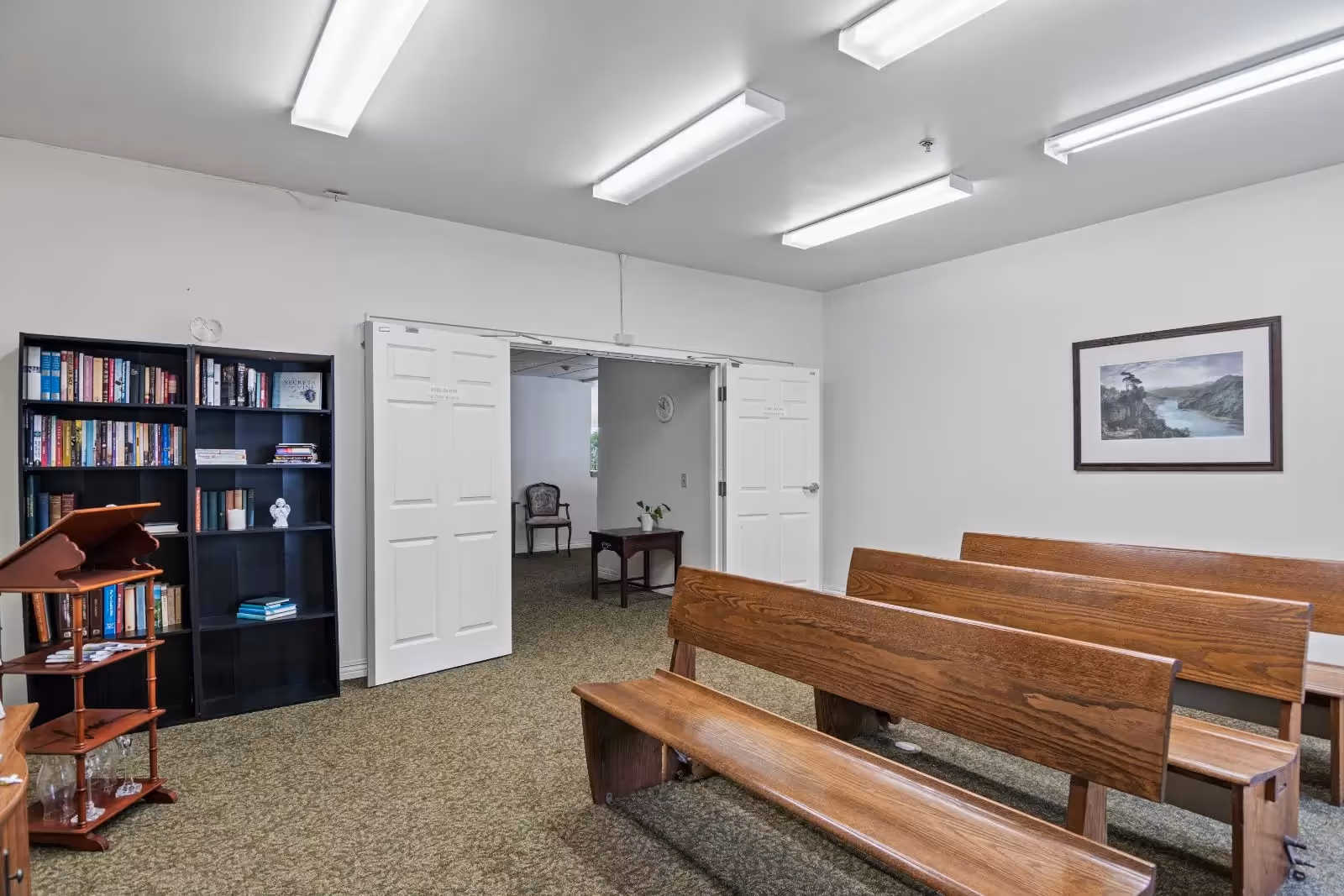 A quiet room with wooden benches arranged in rows, a black bookshelf filled with books, a wooden stand with pamphlets, and a framed landscape picture on the wall. The room has white walls, fluorescent ceiling lights, and an open double door leading to another room with a chair and table.