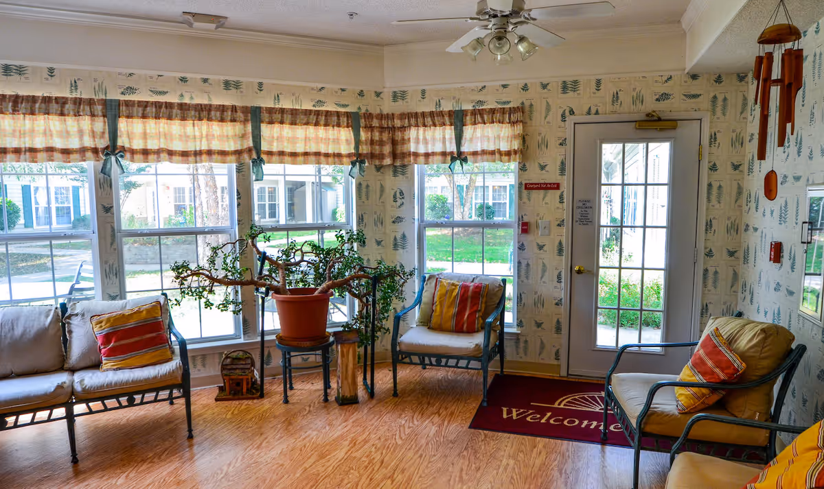 A cozy sitting area with three cushioned chairs featuring colorful striped pillows, a potted plant on a small table, large windows with plaid valances letting in natural light, and a glass door with a 'Welcome' mat on the wooden floor. The walls have wallpaper with a nature-themed pattern and a wooden wind chime hanging near the door.