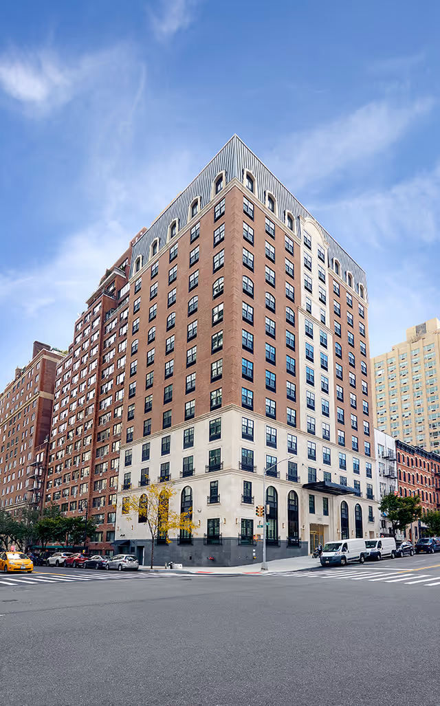 Exterior view of a multi-story brick and stone building on a city street corner under a blue sky with some clouds. Several cars and vans are parked along the street, and a few trees are visible near the building.