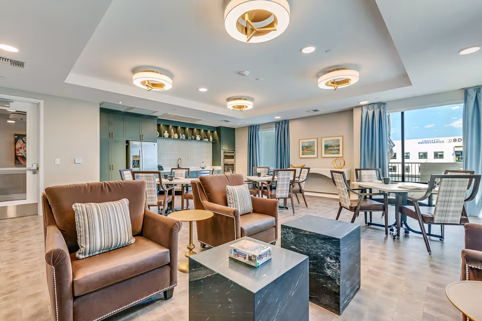 Bright communal lounge with upholstered armchairs, dining tables, and a kitchenette along the back wall.