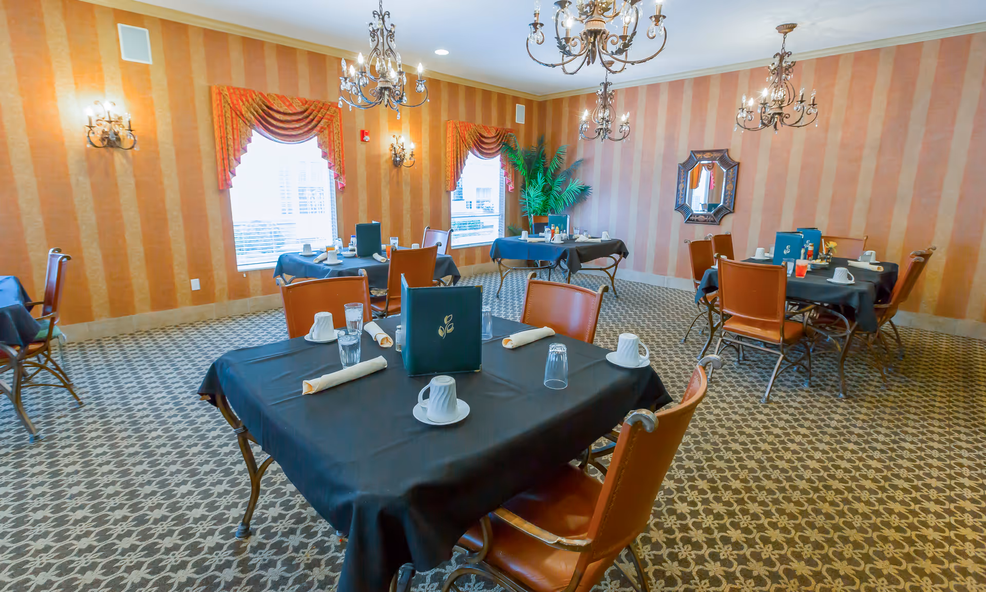 A dining room with several tables covered in black tablecloths, each set with white cups, glasses, and napkins. The room has patterned carpet, striped wallpaper in warm tones, ornate chandeliers, wall sconces, and windows with decorative valances. There is a large green plant in the corner and a decorative mirror on the wall.
