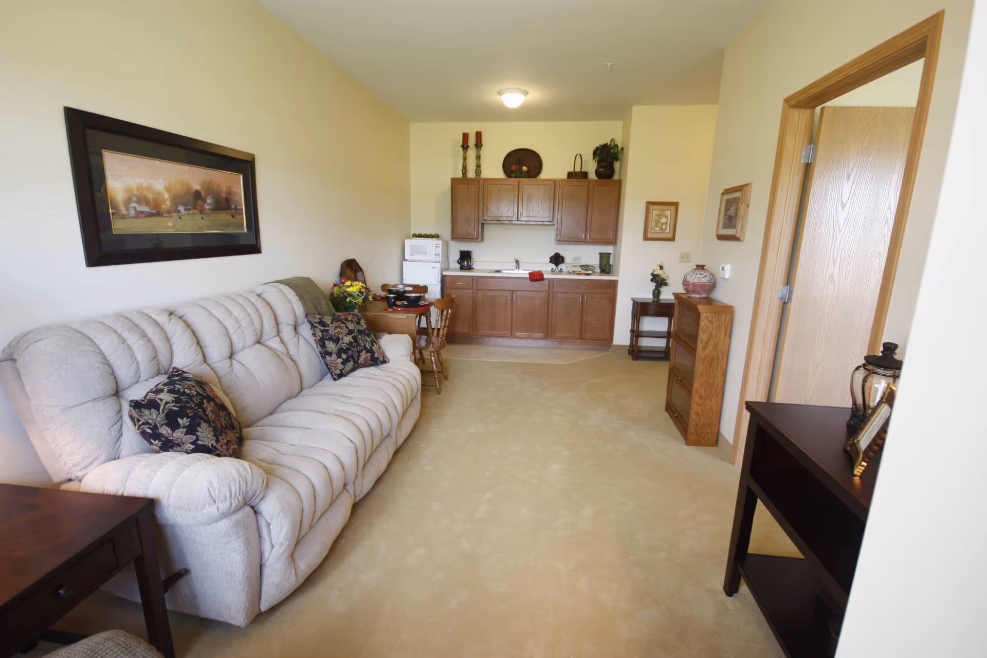 A cozy living area with a beige cushioned sofa adorned with floral pillows, a small wooden dining table with chairs, and a kitchenette with wooden cabinets, a microwave, and a coffee maker. The room has light-colored walls and carpet, with framed artwork and decorative items on the walls and furniture.