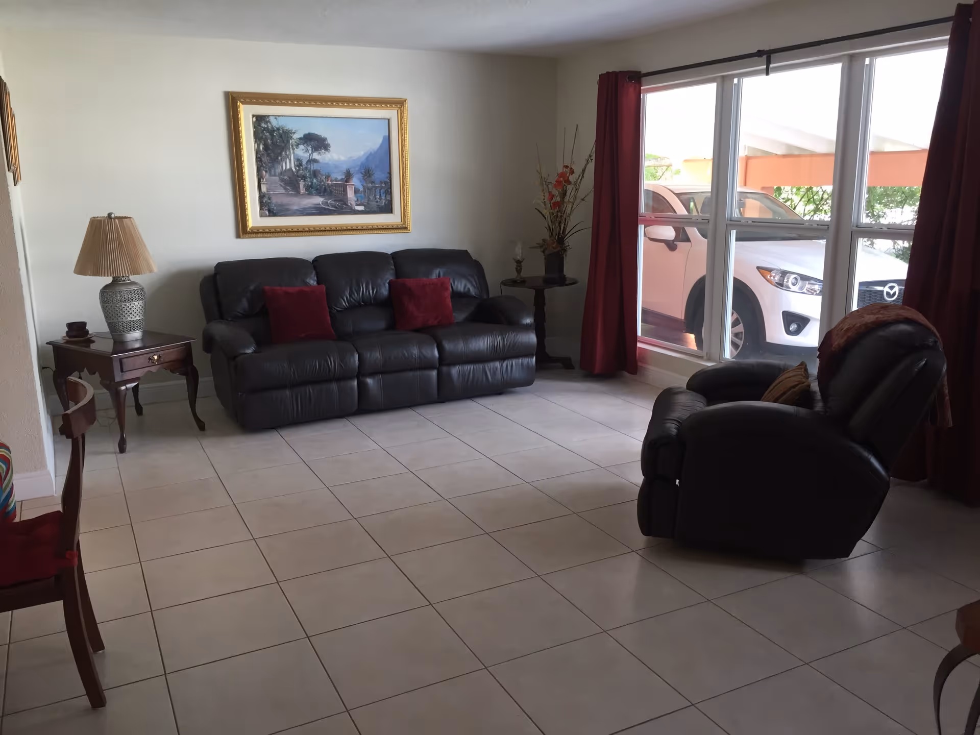 A living room with a black leather sofa and matching recliner chair, both with red and brown cushions. There is a wooden side table with a lamp and a small decorative item next to the sofa. A large window with red curtains shows a white car parked outside. A framed landscape painting hangs on the wall above the sofa, and a tall vase with decorative branches is placed near the window.