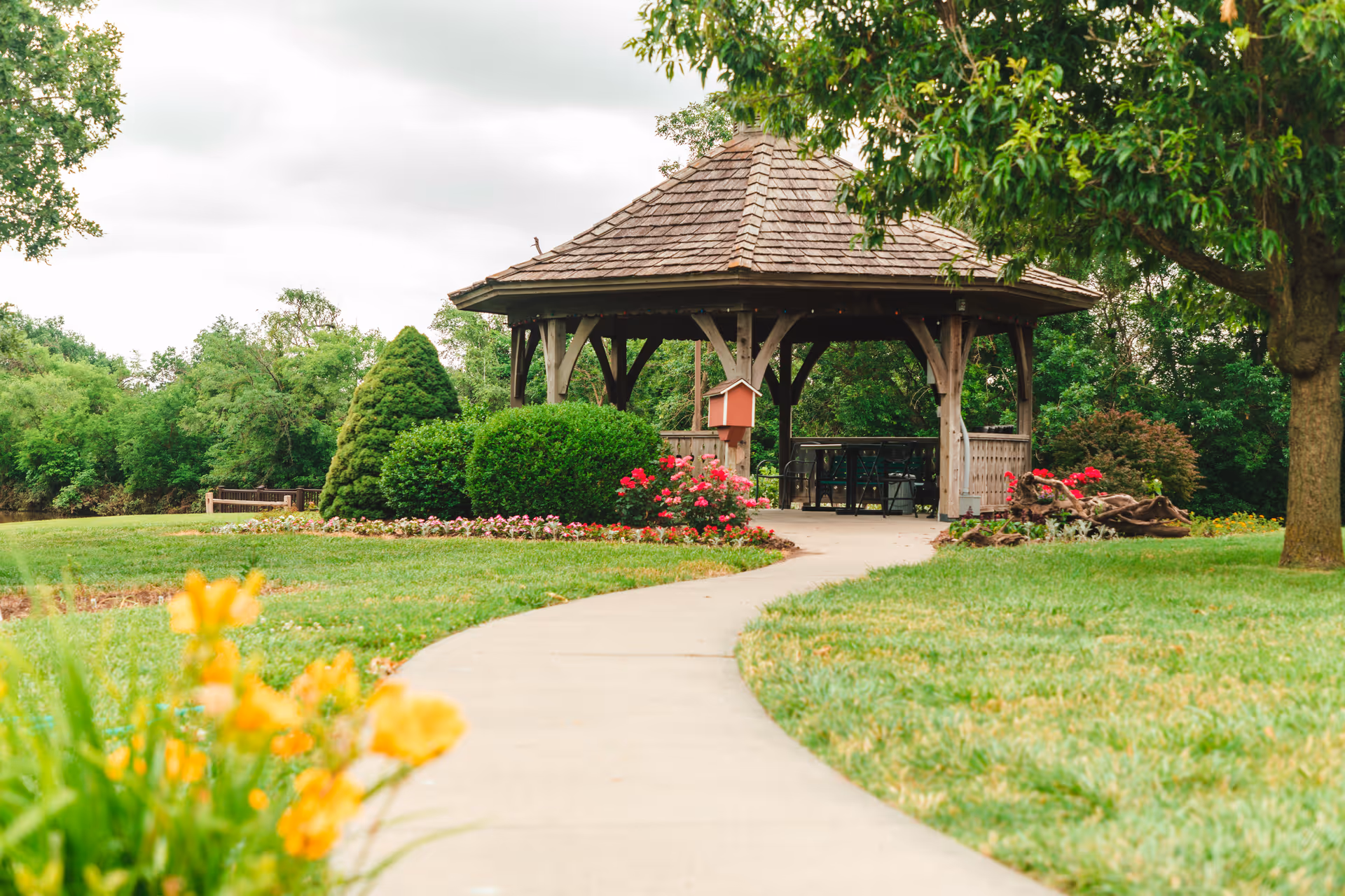 A curved concrete pathway leads to a wooden gazebo surrounded by well-maintained green grass, bushes, and colorful flowers in an outdoor garden setting.