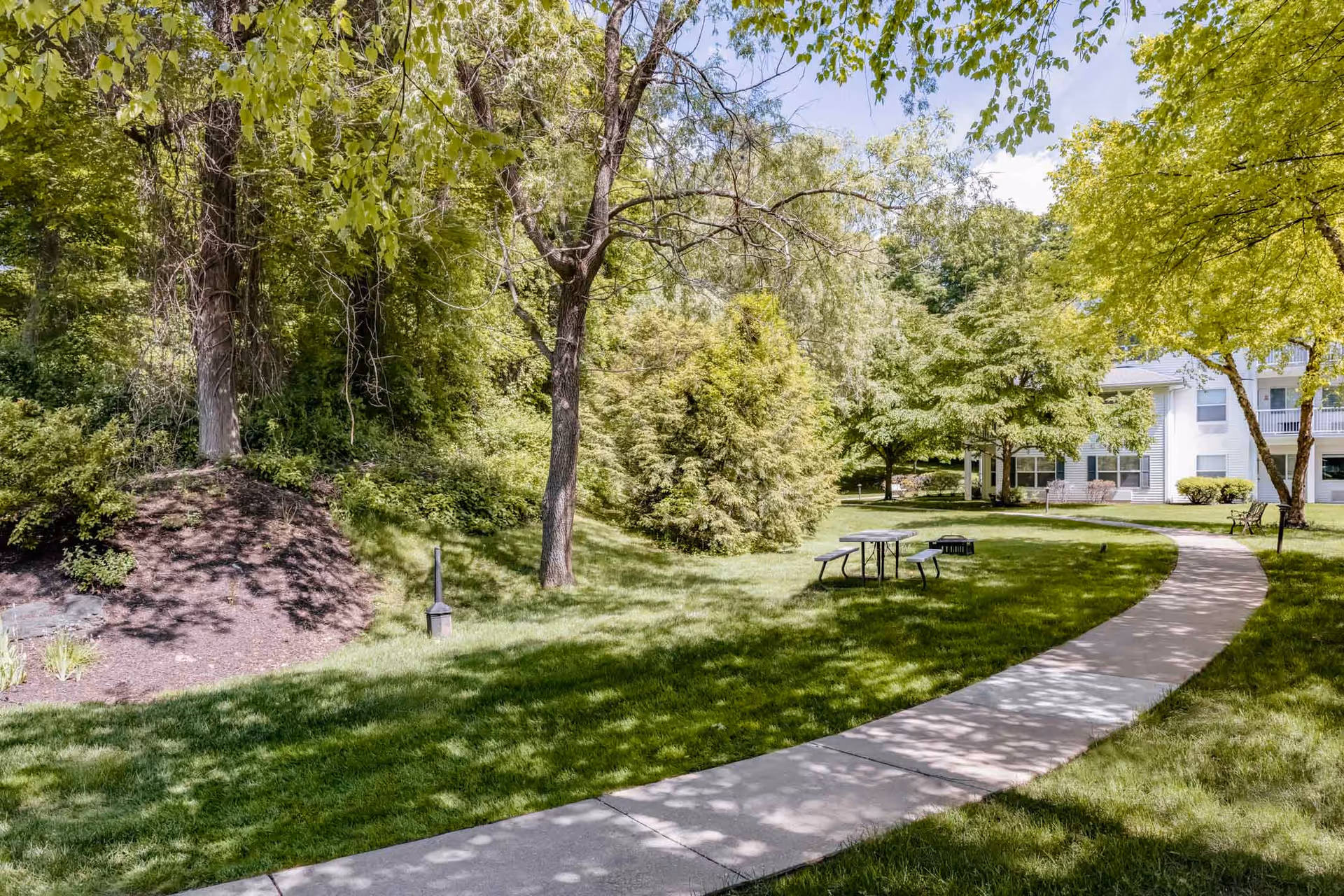 A winding concrete pathway through a green lawn with trees and bushes on either side, leading to a white building with balconies. There is a picnic table and a bench along the path under the shade of the trees.