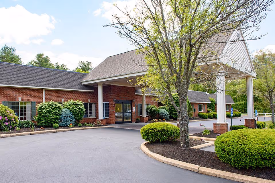 Front exterior view of a single-story brick building with a covered entrance supported by white columns. The driveway curves around landscaped areas with green bushes and a tree with budding leaves. The sky is partly cloudy with blue patches.