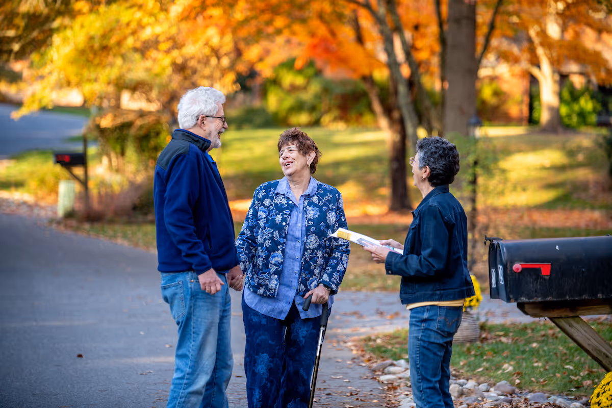 Three elderly people standing and talking on a sidewalk near a mailbox with autumn trees and fallen leaves in the background.