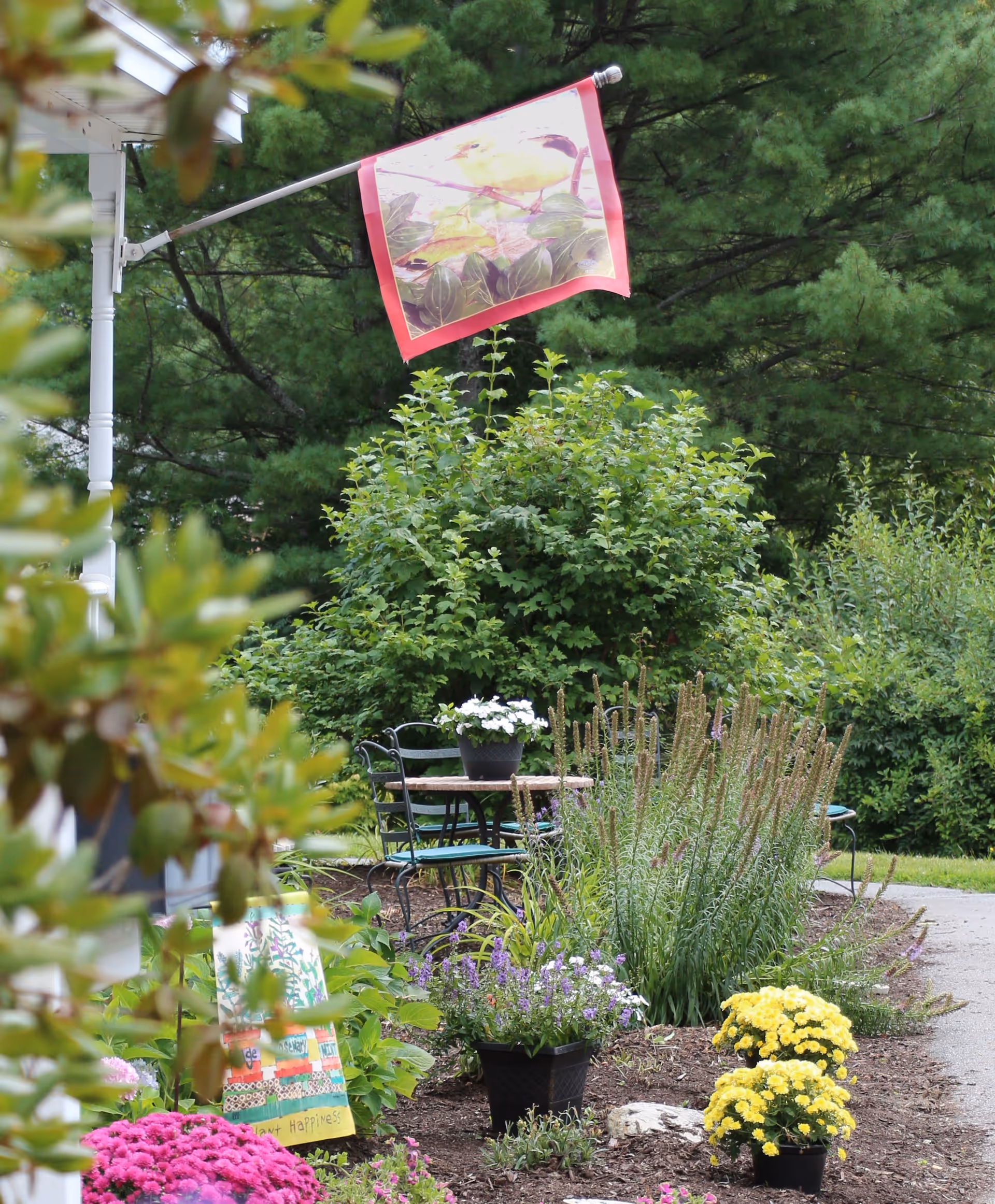 A garden area with various flowering plants including pink, yellow, and purple flowers. There is a small round table with chairs and a potted plant on it. A decorative flag with a bird design is hanging from a white pole attached to a building. The background features dense green trees and bushes.