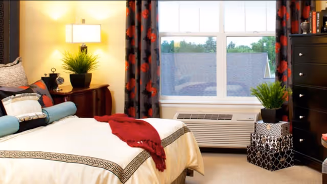 A cozy bedroom with a neatly made bed featuring decorative pillows and a red throw blanket. There is a wooden nightstand with a lamp and a potted plant beside the bed. A window with floral curtains lets in natural light, and below it is an air conditioning unit. Next to the window are two decorative storage boxes with another potted plant on top. A dark wooden dresser is visible on the right side of the room.