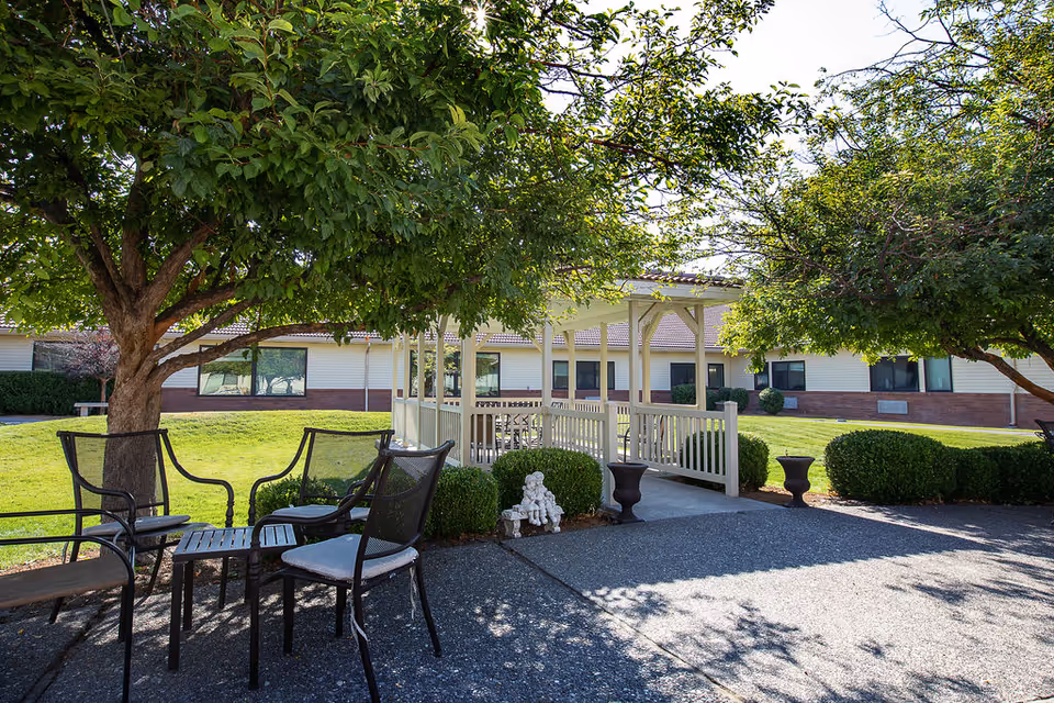 Outdoor seating area with metal chairs and a small table under a large leafy tree. In the background, there is a white gazebo with a railing, surrounded by neatly trimmed bushes and a well-maintained lawn. The building of the facility is visible behind the gazebo under a clear sky.
