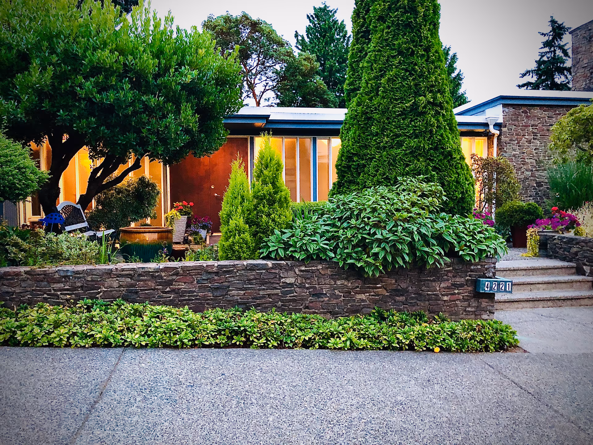 Front view of a single-story house with a stone planter wall, steps, and lush landscaping in front of illuminated windows.
