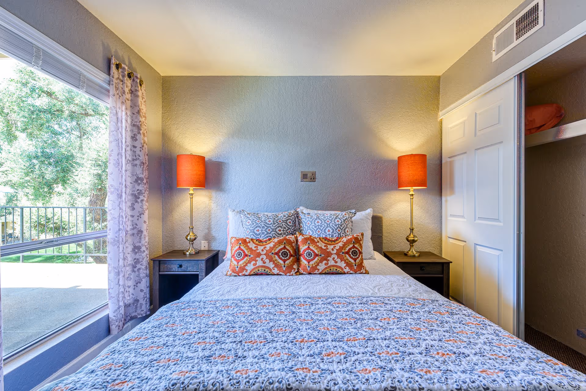 A cozy bedroom with a bed centered against a light gray textured wall. The bed is made with a patterned quilt and decorative pillows in orange and blue tones. On each side of the bed are dark wooden nightstands with brass lamps featuring orange lampshades. To the left, there is a large window with sheer floral curtains, letting in natural light and showing greenery outside. On the right side, there is a closet with white sliding doors, one door partially open revealing shelves with folded items.