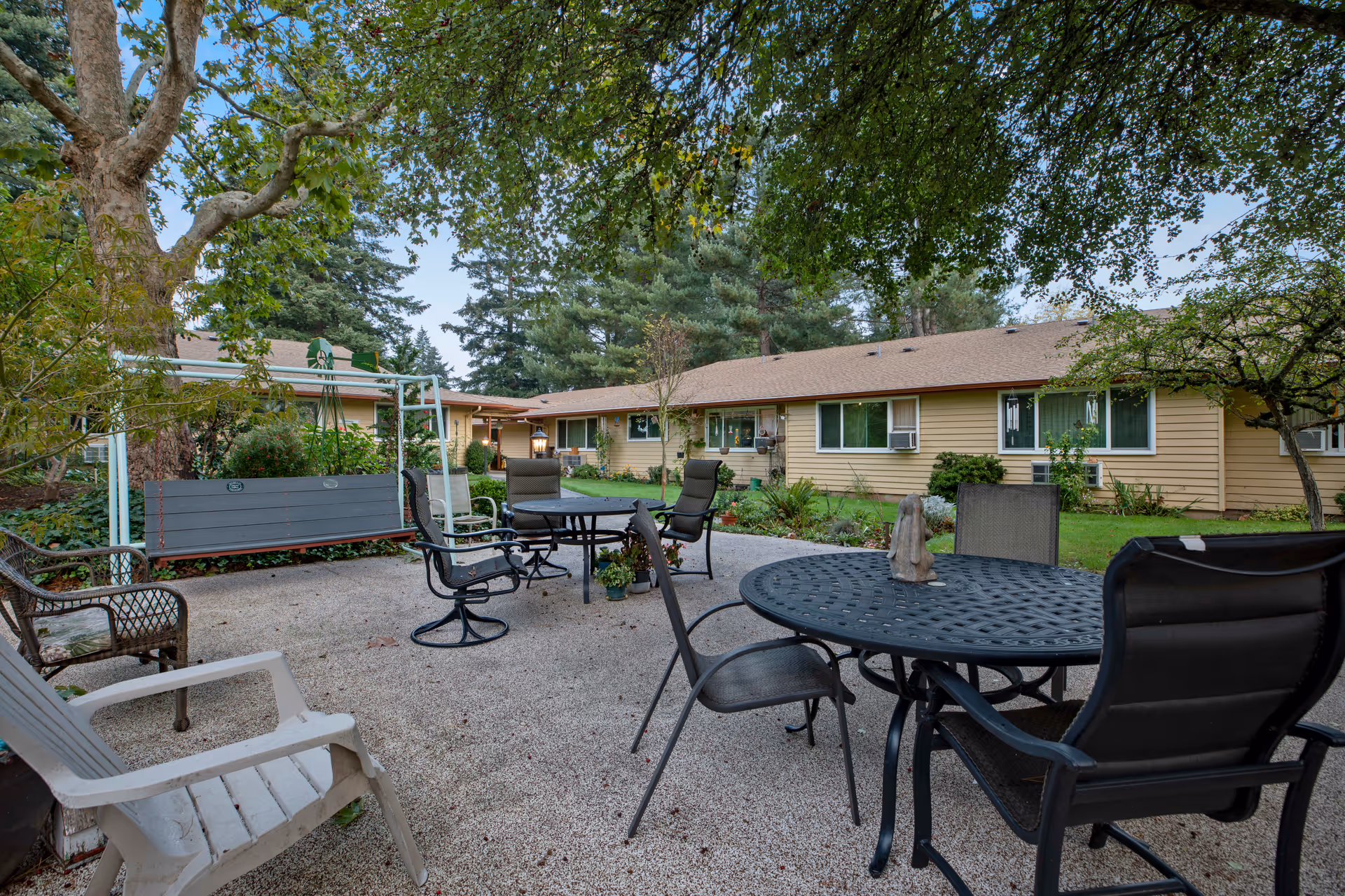 Outdoor patio area at Parkview Christian Retirement Community with multiple seating options including metal tables and chairs, a wooden swing, and plastic chairs. The patio is surrounded by trees and greenery, with a single-story beige building in the background under a partly cloudy sky.
