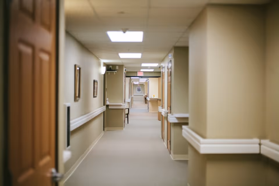 A long, well-lit hallway in a senior living facility with beige walls, carpeted floor, wooden doors on the right side, handrails along the walls, and framed pictures hanging on the left wall.