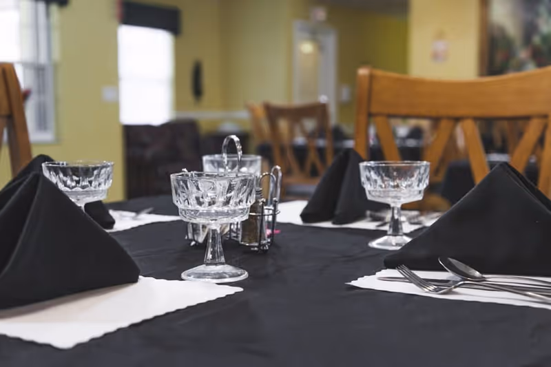 A dining table set with black cloth napkins folded in a triangular shape, crystal dessert bowls, silverware, and a small condiment holder in the center. Wooden chairs surround the table, and the background shows a softly lit room with yellow walls and windows.
