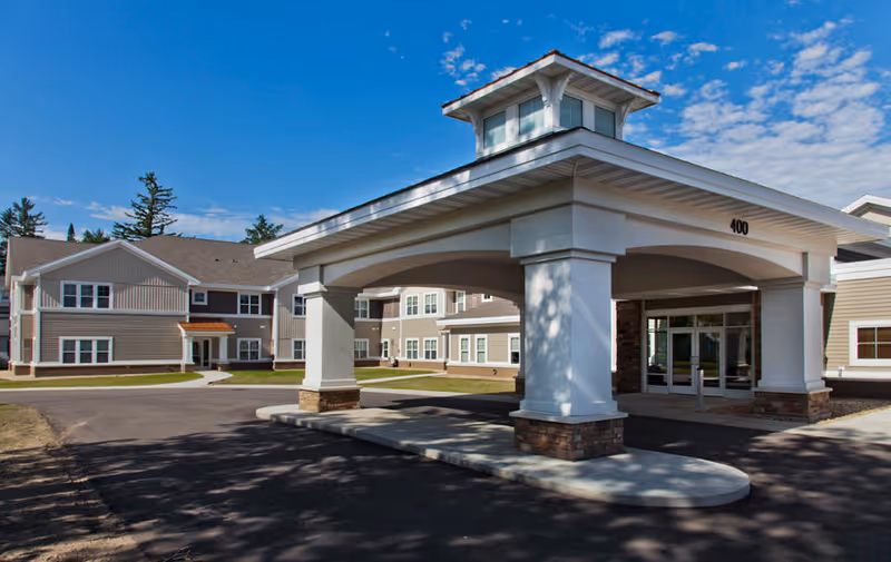Exterior view of St. Mark's Living assisted living and memory care facility showing a large covered entrance with white pillars and a beige building with multiple windows under a blue sky with some clouds.