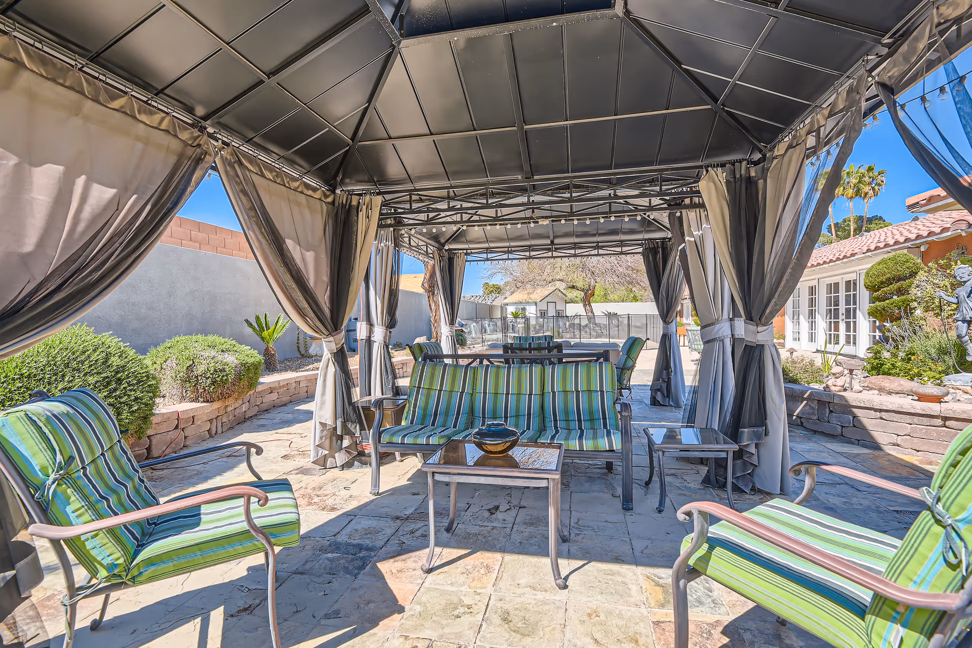 Outdoor patio area with a metal-framed gazebo covered by a dark canopy. Inside the gazebo are cushioned chairs and a loveseat with green and blue striped cushions, along with glass-top tables. The patio is paved with stone tiles and surrounded by landscaped bushes and trees. A building with white-framed windows and a tiled roof is visible in the background under a clear blue sky.