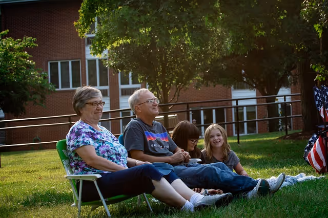Two elderly adults and two children sitting on the grass outside near a building, enjoying a sunny day with trees and greenery around them.