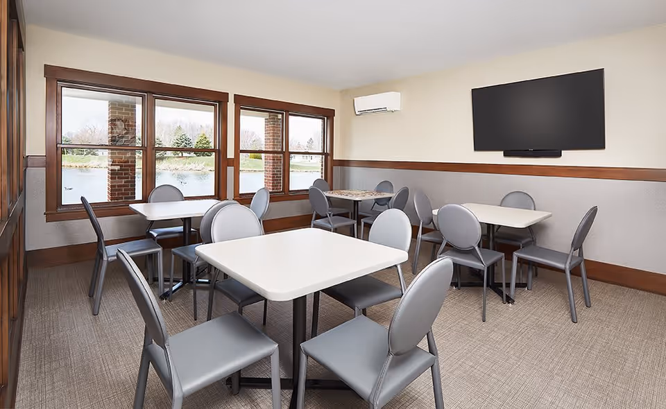 Small dining/activity room with several square tables, gray chairs, large windows overlooking a pond, and a wall-mounted TV.