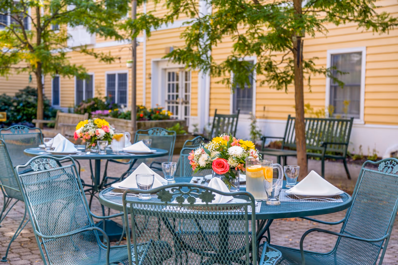 Outdoor patio area with round metal tables and chairs set for dining. Each table has a floral centerpiece, glasses, napkins, and a pitcher of water with lemon slices. The background shows a yellow building with white trim, windows, and some greenery including trees and flower beds.