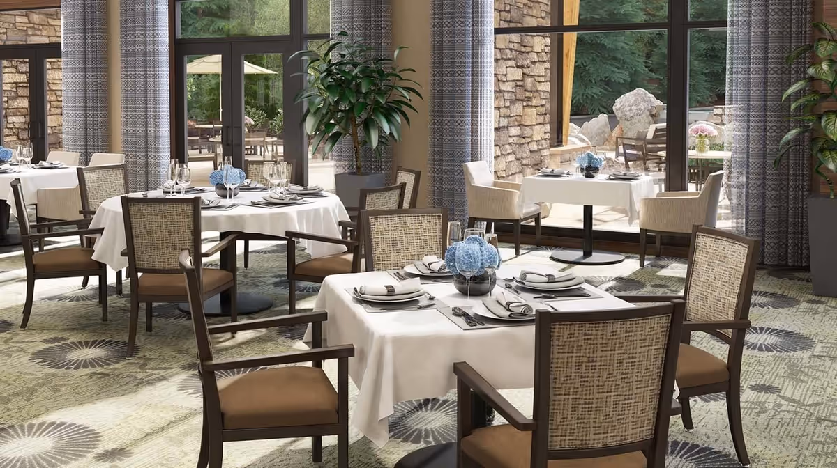 A bright dining room with round and square tables covered with white tablecloths, set with plates, napkins, glasses, and silverware. Each table has a centerpiece with blue flowers. The room has large windows with patterned curtains, allowing natural light and a view of an outdoor patio area with stone walls and greenery. There are cushioned chairs with wooden frames around the tables and potted plants near the windows.