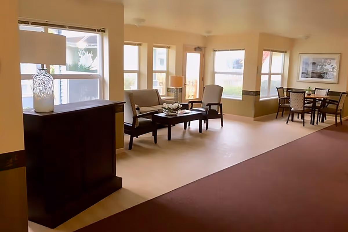 A bright and spacious common area in a senior living facility with large windows letting in natural light. The room features a seating area with a loveseat, two armchairs, and a coffee table with a flower arrangement. There is a dark wooden cabinet with a decorative lamp on top near the windows. In the background, there is a dining area with a round table and four chairs, and a framed picture on the wall.
