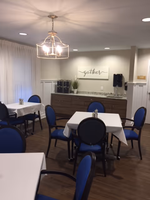 A dining room with several tables covered with white tablecloths and surrounded by blue cushioned chairs. A modern chandelier hangs from the ceiling. In the background, there is a countertop with a coffee machine, a small plant, and a wall decoration with the word 'gather'. The room has wood flooring and light-colored walls.