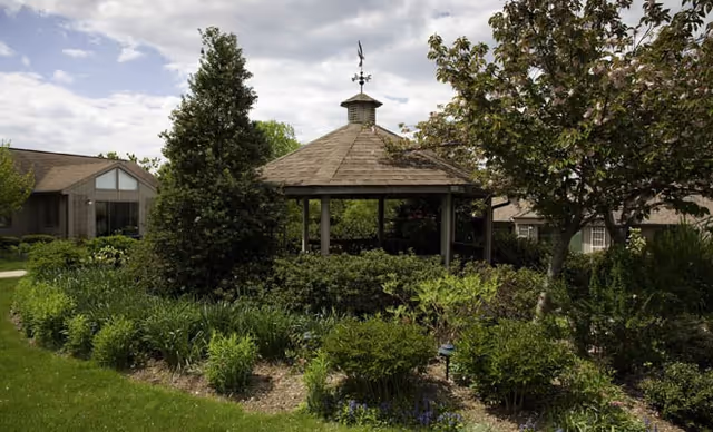 A garden area with a wooden gazebo surrounded by various green shrubs, trees, and flowering plants under a partly cloudy sky. Residential buildings are visible in the background.
