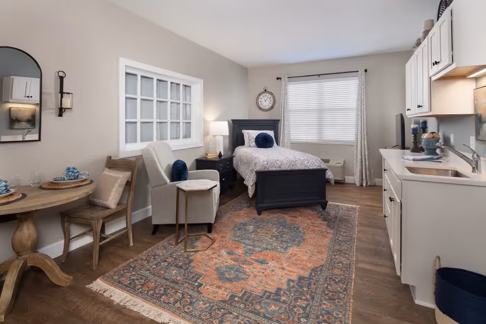 A cozy studio apartment featuring a single bed with patterned bedding and a blue round pillow, a wooden nightstand with a lamp, a large window with curtains, a comfortable armchair with a small side table, a round wooden dining table with two chairs set with plates and glasses, a kitchen area with white cabinets, a sink, and a countertop, and a colorful patterned rug covering the wooden floor.