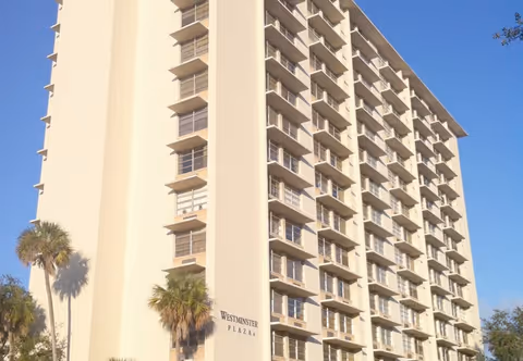 Exterior view of a tall, multi-story beige building named Westminster Plaza with balconies on each floor and palm trees in front under a clear blue sky.
