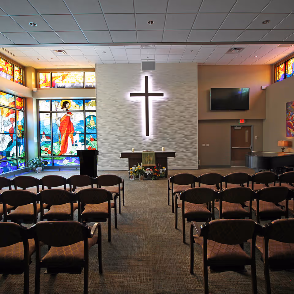 Interior of a chapel with rows of chairs facing a wall with a large illuminated cross. To the left, there are colorful stained glass windows depicting religious imagery. In front of the cross is a small altar with flowers and candles. A piano is visible on the right side of the room.