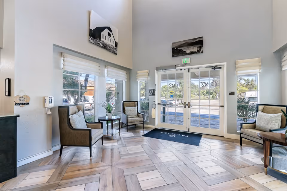 A bright and welcoming senior living facility lobby with large windows and glass double doors letting in natural light. The room features light-colored walls, wood-patterned tile flooring, three cushioned armchairs with pillows, small round tables with plants, and framed black-and-white photographs on the walls.