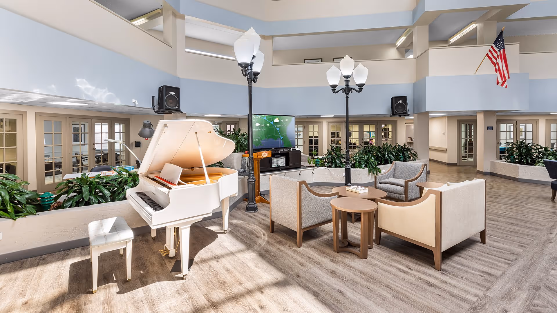 A bright and spacious common area in a senior living facility featuring a white grand piano with a matching bench, several comfortable armchairs arranged around small wooden tables, two tall streetlamp-style light fixtures, a large flat-screen TV, and various green plants. The room has light wood flooring, large windows, and an American flag displayed on the wall.