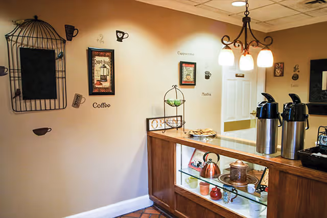 A small coffee station with two stainless coffee urns on a wooden counter, decorative teapots and mugs on glass shelves, and coffee-themed wall art and lighting.
