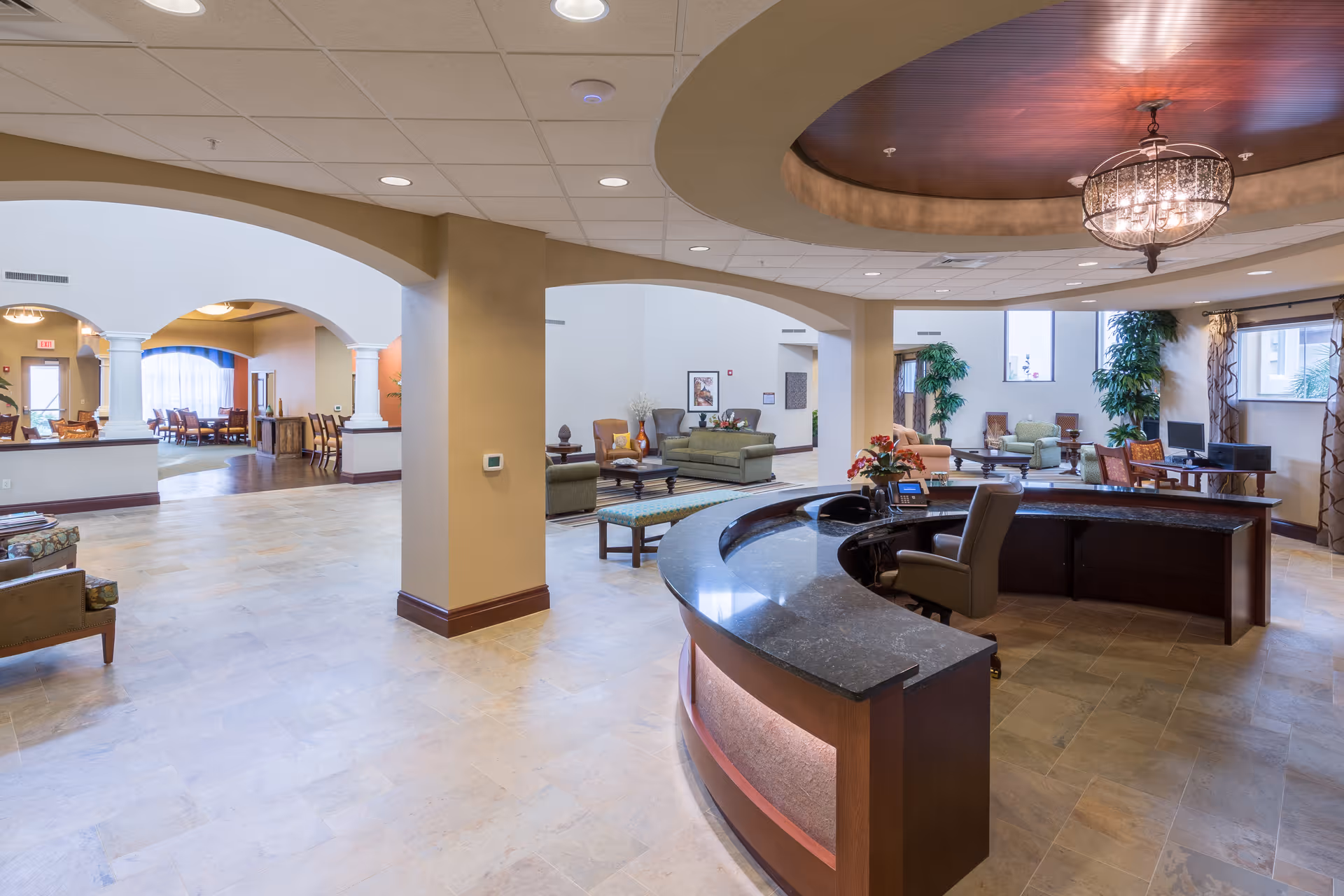 Spacious senior living facility interior with a curved reception desk in the foreground, comfortable seating areas with sofas and chairs, large windows letting in natural light, decorative plants, and an elegant chandelier hanging from a recessed ceiling.