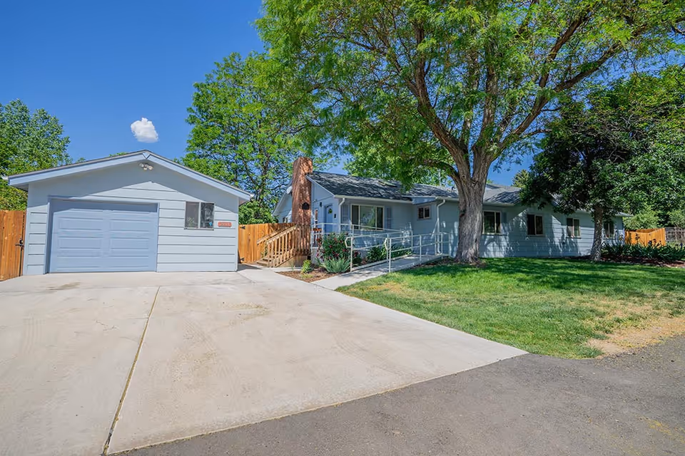Exterior view of a single-story assisted living facility with light blue siding, a large tree in the front yard, a concrete driveway leading to a garage, and a wooden fence surrounding the property under a clear blue sky.