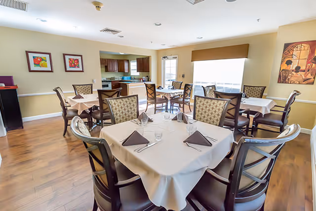 A bright dining room with several tables covered in white tablecloths, each set with folded brown napkins, glasses, and silverware. The room features wooden chairs with patterned cushions, hardwood flooring, and light yellow walls adorned with framed artwork. In the background, there is a kitchen area with wooden cabinets and a door leading outside.