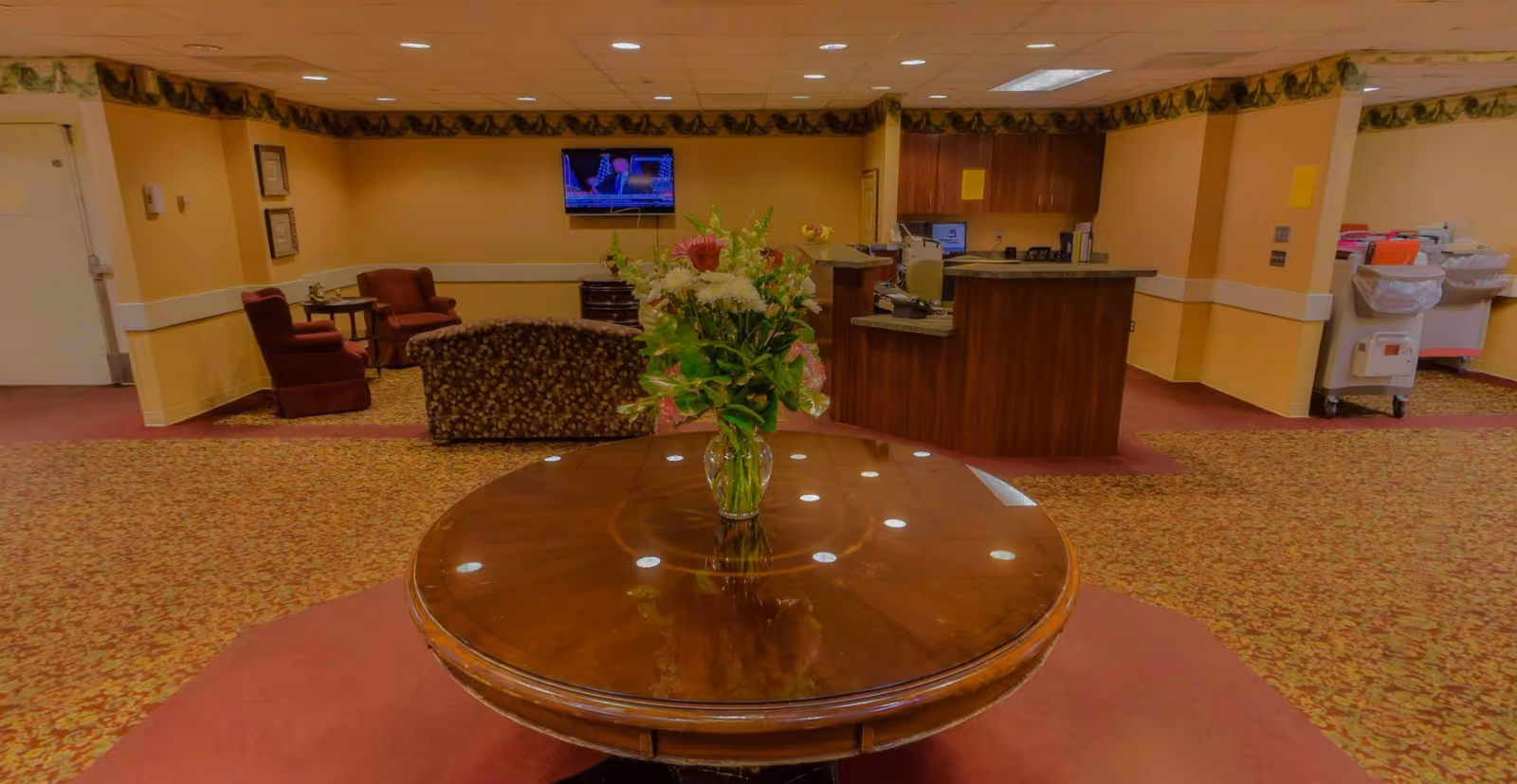 Interior view of a senior living facility lobby area with a round wooden table in the foreground holding a vase of flowers. In the background, there is a reception desk, a wall-mounted TV, and a seating area with armchairs and a sofa. The walls are painted yellow with a decorative border near the ceiling, and the floor is carpeted with a patterned design.