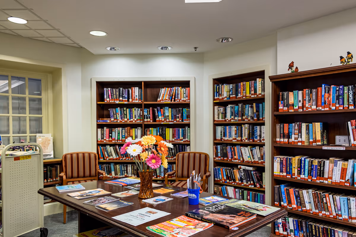 A cozy library room with wooden bookshelves filled with books along the walls. A wooden table in the center holds magazines, brochures, a vase with colorful flowers, and a cup with pens. Two striped armchairs are positioned near the table, and a book cart is visible near a window with multiple panes.