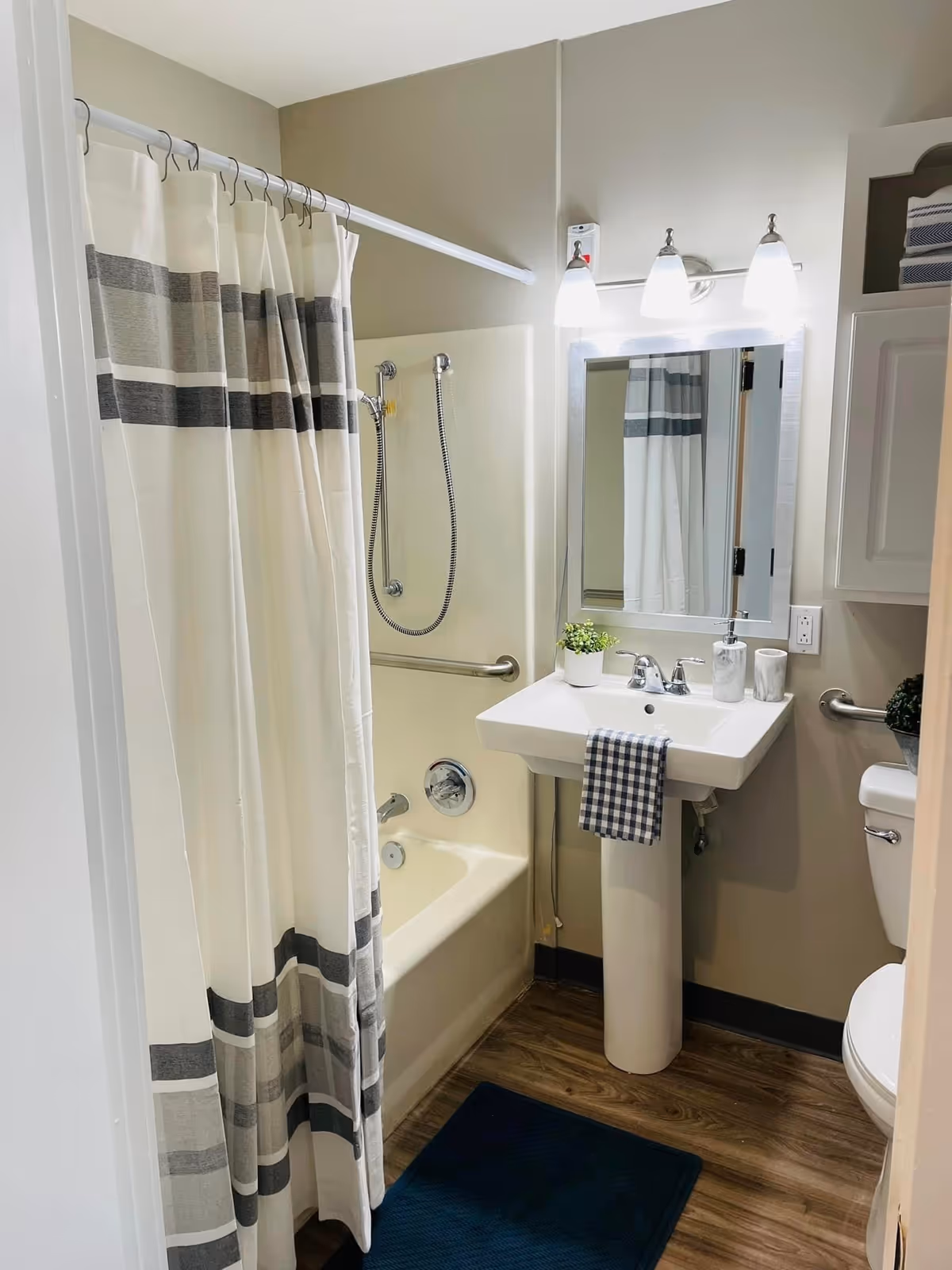 A small bathroom with a bathtub and shower combination featuring a striped shower curtain. There is a white pedestal sink with a checkered hand towel hanging on it, a mirror above the sink with three light fixtures, a toilet partially visible on the right, and a cabinet with folded towels. The floor has wood-like vinyl flooring and a dark blue bath mat.