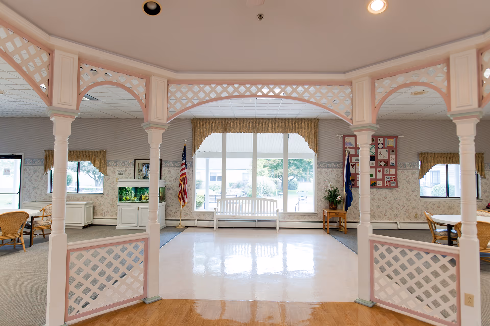 Interior view of a common area in a senior living facility featuring white lattice archways, a white bench in front of large windows with beige curtains, an American flag, a fish tank, and wicker chairs with tables on either side.