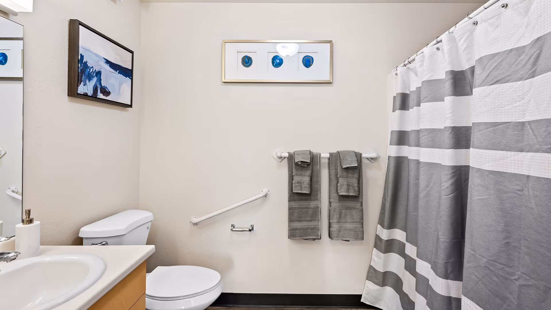 A clean bathroom featuring a white toilet, a sink with a soap dispenser, a large mirror, and a shower with a gray and white striped curtain. Two gray towels hang on a towel rack on the wall, which also has two framed abstract artworks.