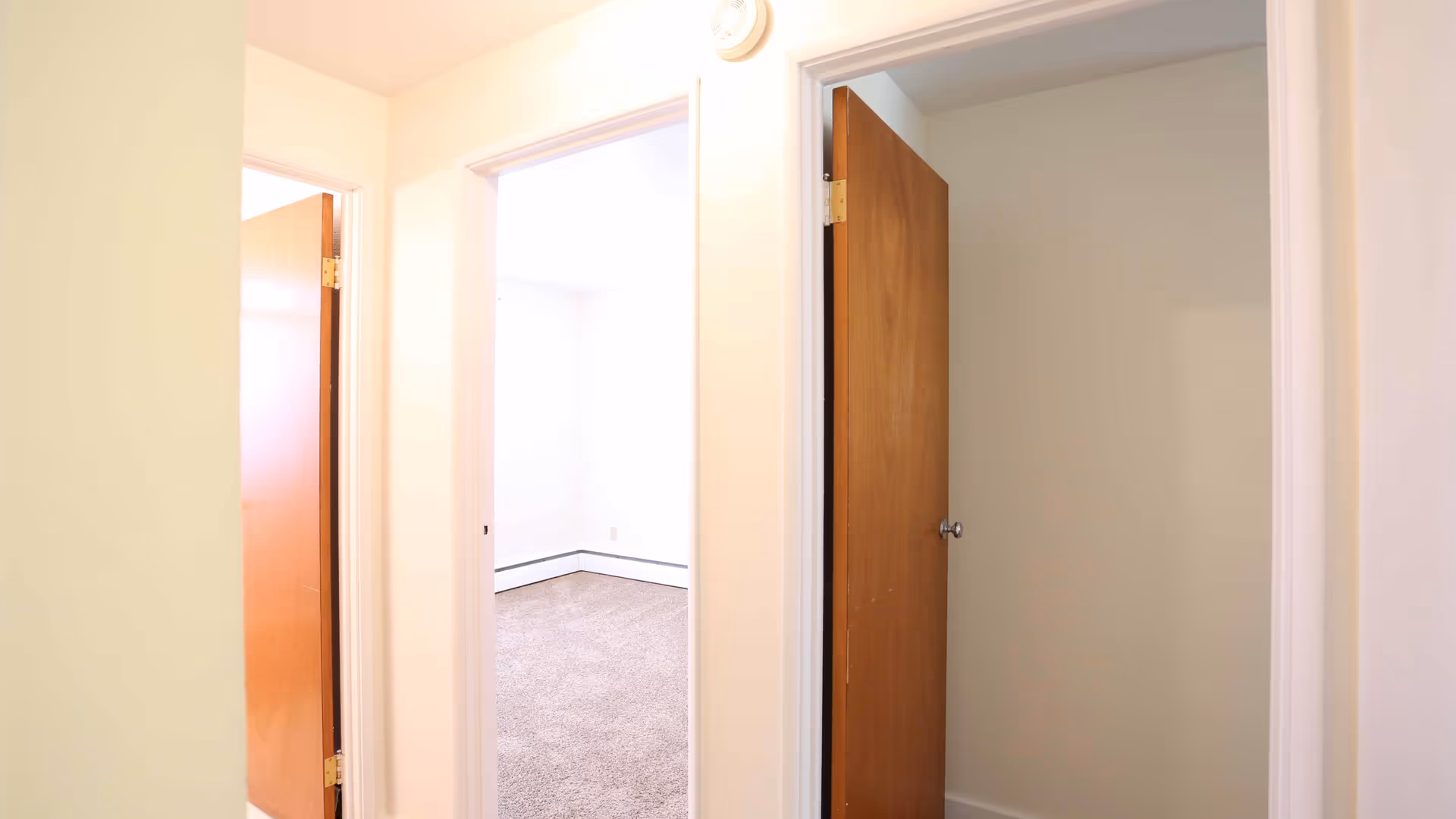 Bright interior hallway with two open wooden doors revealing an empty carpeted room and a small closet.