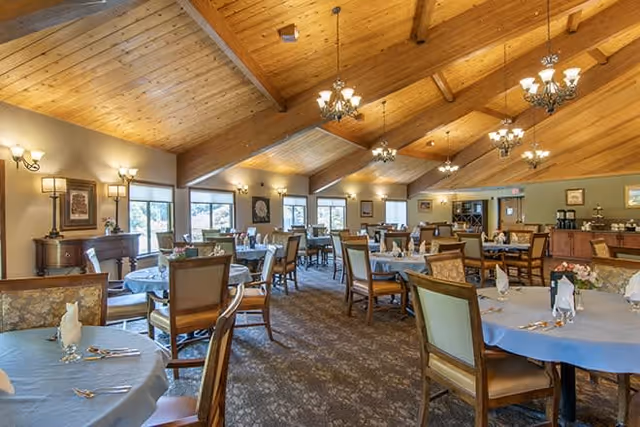 Spacious dining room with wooden vaulted ceiling and chandeliers, multiple round tables covered with light blue tablecloths, each set with napkins and silverware, surrounded by wooden chairs with cushioned seats. Large windows let in natural light, and the walls are decorated with framed pictures and sconces.