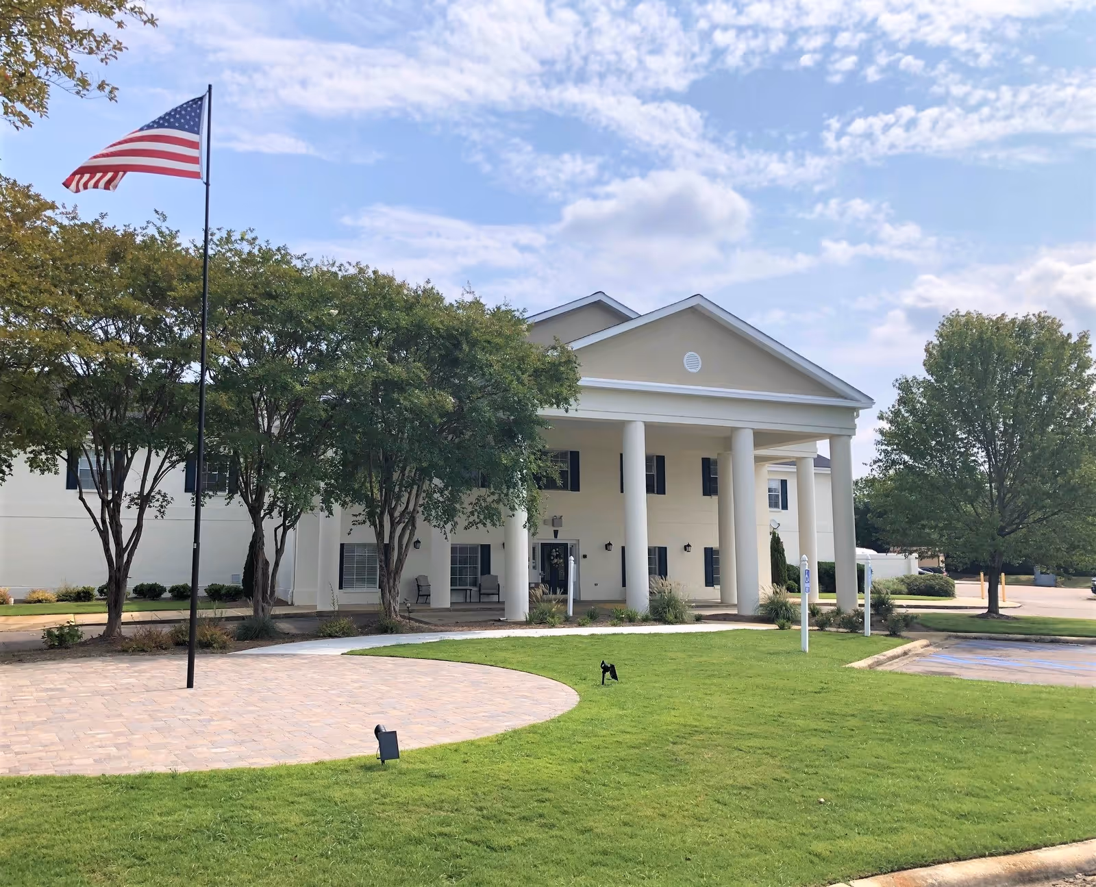 Front exterior view of a two-story building with white columns and a triangular pediment, surrounded by green trees and a well-maintained lawn. An American flag is flying on a flagpole in front of the building under a partly cloudy sky.