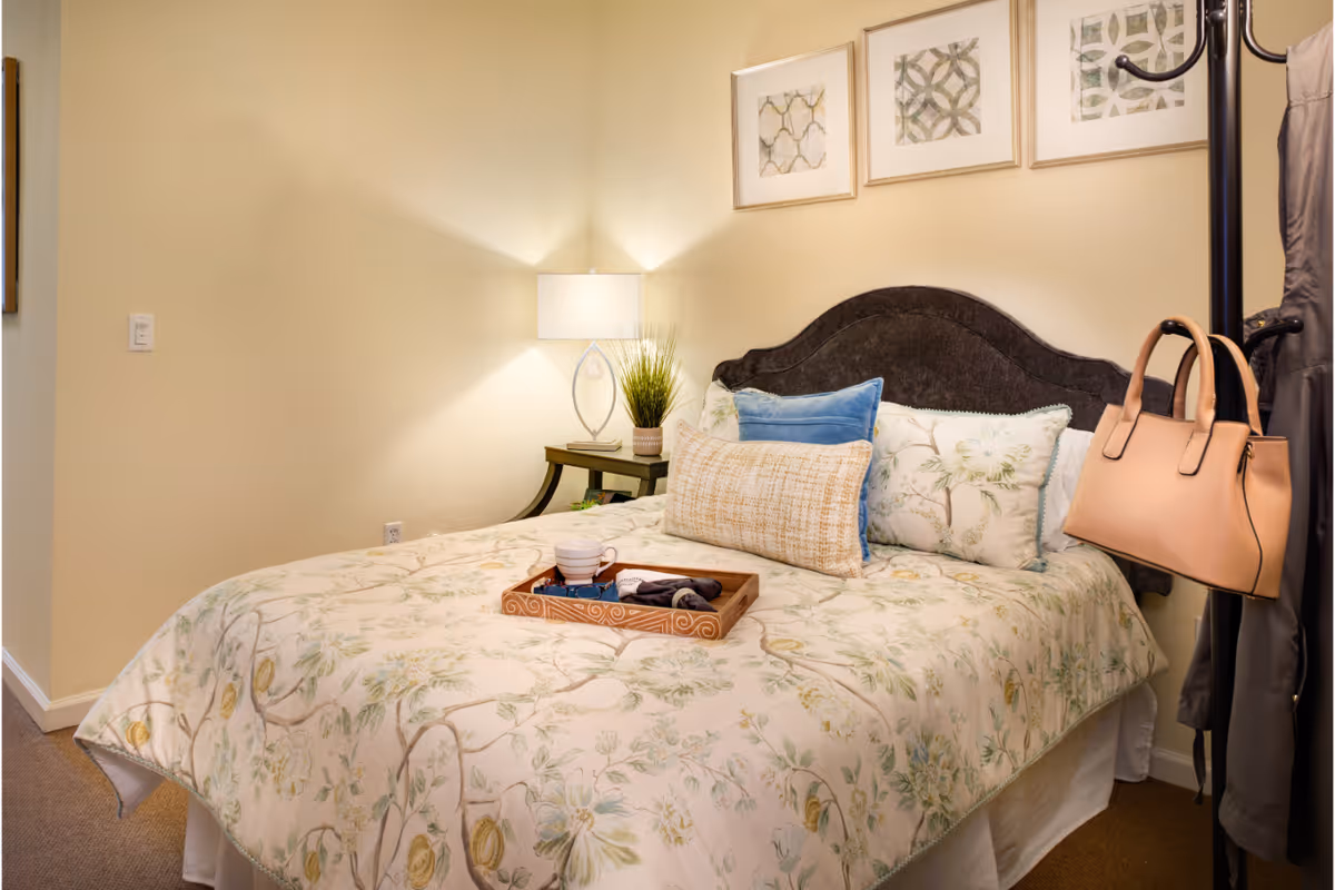 A cozy bedroom with a floral patterned bedspread on a bed with a dark upholstered headboard. The bed has several pillows including a blue one and a rectangular beige one. A wooden tray with a cup and some items is placed on the bed. There is a nightstand with a modern lamp and a small plant. Three framed abstract artworks hang on the wall above the bed. A coat rack with a beige handbag and a gray coat is visible on the right side.