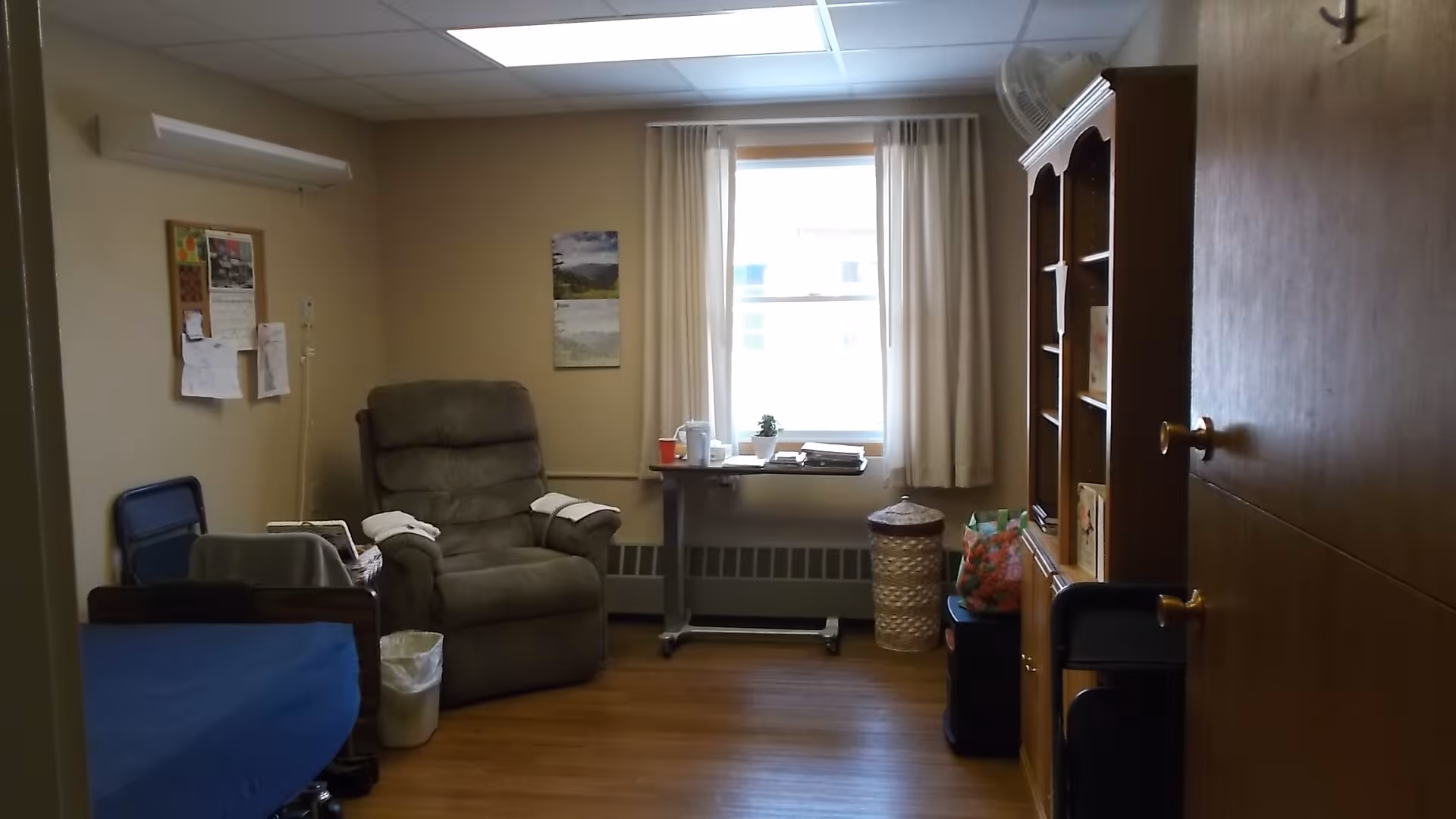 A small room in a senior living facility with a recliner chair, a hospital-style bed with blue bedding, a small table with items on it in front of a window with white curtains, a wooden cabinet with shelves, and a bulletin board on the wall.